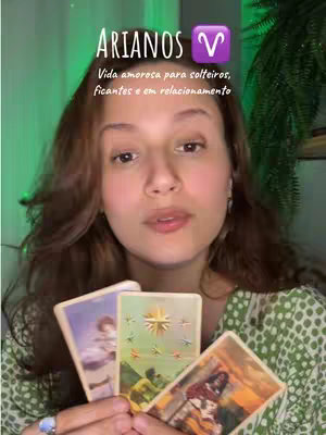 A woman with long brown hair is centered in the frame, holding three tarot cards. Above her, text overlaid says 'ARIANOS' with an Aries symbol. The woman is wearing a green patterned top. The background is blurred but appears to be a room with green and a plant visible on the right side.