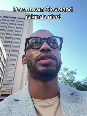 A man wearing glasses and a blazer looks up at the camera with a tall building visible behind him. Overlaid at the top is text reading, "Downtown Cleveland is kinda nice!"