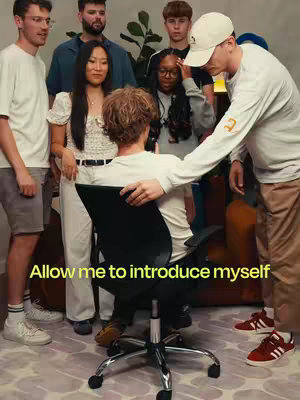 A group of young adults are gathered in a room. A person is seated in a black office chair and is being introduced by one of the group members. The hook in the image is "Allow me to introduce myself".