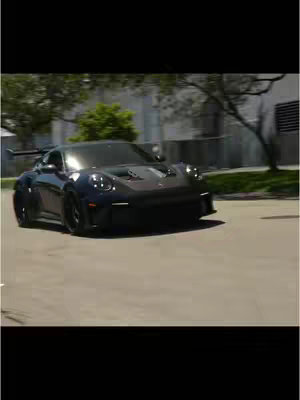 The cover image features a sleek, dark blue Porsche sports car, captured in motion on a paved road. The car is the central focus, positioned at a slight angle, implying movement. The background includes blurred trees and a building, suggesting an outdoor setting. The composition is well-lit, with the car's body reflecting the ambient light.