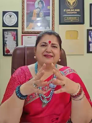 A woman with a red bindi on her forehead sits facing the camera. She is wearing a red saree and multiple beaded necklaces and bracelets. Behind her are framed pictures and awards.