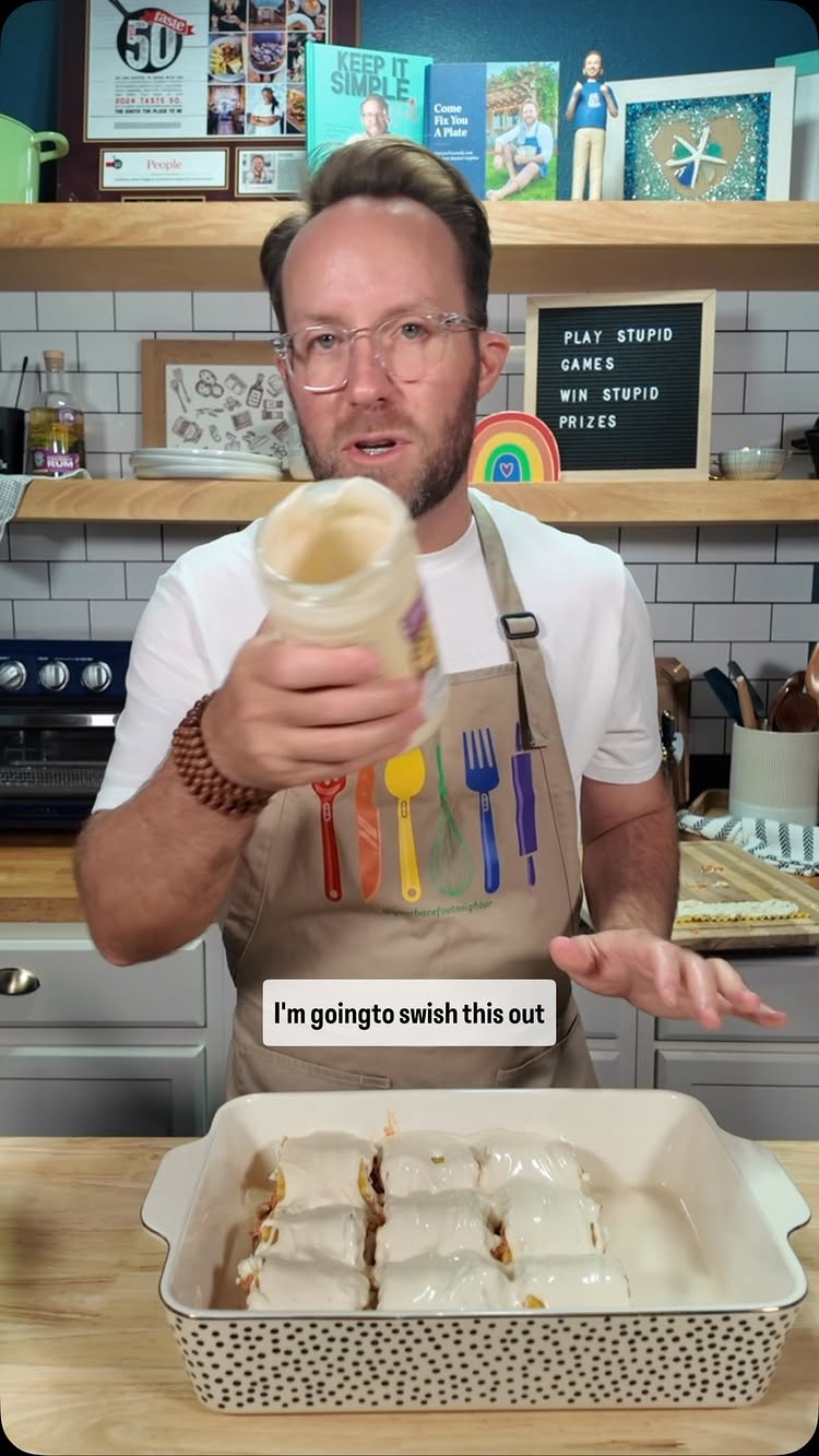A man in a white shirt and apron is holding a jar of white sauce over a casserole dish filled with food that is covered in white sauce. The scene is set in a kitchen with cabinets, a shelf with various items and a sign that reads “PLAY STUPID GAMES WIN STUPID PRIZES”.