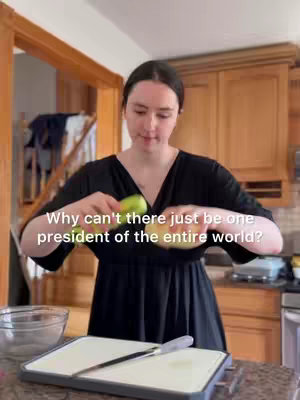 The cover image features a woman in a kitchen holding a green apple and a piece of bread. The image is brightly lit, and the woman is the central focus, with kitchen counters and cabinets providing the background. A text overlay stating 'Why can't there just be one president of the entire world?' is displayed on the image.