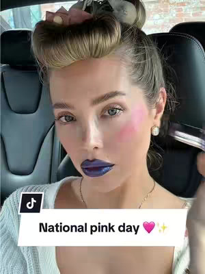 A selfie-style shot featuring a woman with makeup in a car. She has blonde hair styled up with a hair accessory. Her face is the focus, with dark blue lipstick and pink blush. A text overlay at the bottom reads "National pink day". The background is blurred, showing the car interior.