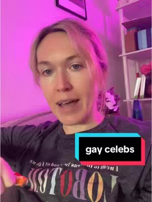 A woman with light-colored hair is filming herself indoors. The background is a bright pink, the lighting appears to be coming from lamps on either side of her. There's a framed picture on the wall and a white shelf with items on it. The woman is wearing a dark t-shirt with a colorful design.