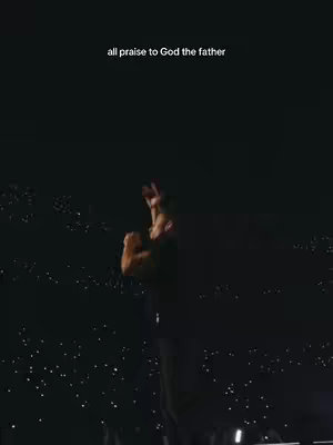 A low-angle shot of a person with their arms raised in a worshipful gesture. The background is dark, suggesting a night setting or a stage, with small, dispersed lights that could be from a crowd or stage lighting. Text overlayed at the top reads 'all praise to God the father'.