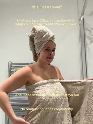 A woman wrapped in a beige towel after a shower is looking at the camera and smiling. There is text overlay at the top that says It's just a towel, and other text overlays in the lower middle and bottom. The background is a light-colored bathroom setting with a towel rack and shower or glass in the back right.