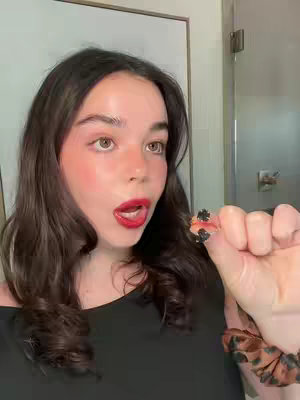 A woman with dark hair and red lipstick holds up a black ring, looking surprised or excited. She is positioned against a plain white wall and a partially visible bathroom door.