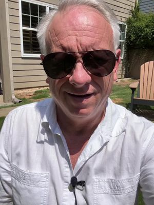 An older white man wearing sunglasses is taking a selfie outside on a sunny day. The man is wearing a white shirt, and behind him a grassy backyard scene is visible with a section of a beige house with a window and a wooden chair.