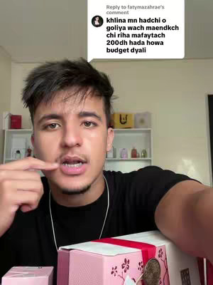 A young man with dark hair and a black t-shirt speaks directly to the camera, gesturing with his finger. Behind him, a room is visible with shelves holding various products, and in the foreground are gift boxes.  A comment box from a user is on the top right of the image.