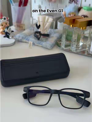 The cover image showcases a white desk with various items arranged on it. In the foreground, there's a pair of black-framed glasses and a black glasses case. Behind these, the background is cluttered, including items like an open case, a clear glass, a pillow with a design, and other decorative objects. The overall composition appears to be a product showcase or a lifestyle shot.
