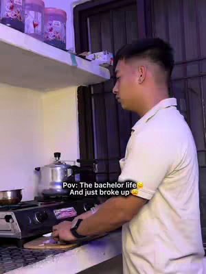 A man is in a kitchen, standing over a stove with a chopping board. He is wearing a white shirt. The kitchen has a light-colored countertop, with shelves above holding jars and other items. The image is shot from a side angle.