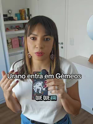 A woman with dark hair and a white top looks towards the camera, holding a small card with a drawing. Text overlays read "Urano entra em Gêmeos" and "QUE QUE VAI". The background includes a bookshelf and a mirror.