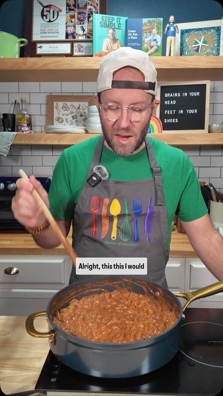 A man in a green shirt and apron is stirring a pot of food on a stovetop. The background includes kitchen elements, cookbooks, and a sign with the phrase "Brains in your head, feet in your shoes". Text overlayed at the bottom: "Alright, this this I would". The composition is well-lit, focusing on the cooking action and the man's expression.