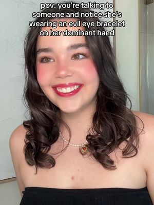 A young woman with dark hair, rosy cheeks, red lipstick, and a pearl necklace is smiling at the camera. Overlayed text reads: "pov: you're talking to someone and notice she's wearing an evil eye bracelet on her dominant hand."