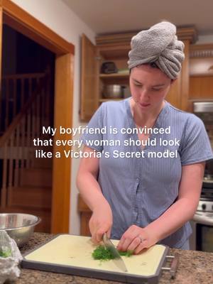 A woman wearing a towel turban is in a kitchen, cutting herbs on a cutting board while looking down. The frame includes kitchen cabinets and a glimpse of a staircase in the background.
