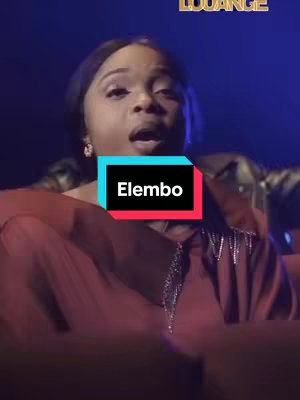 The cover image features a woman in the center, singing with her mouth open. She has dark hair. The background is dark with some blue and purple lighting. There is a text overlay in a rectangle with the text 'Elembo'.