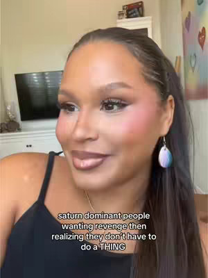 A woman with a dark complexion and dark hair is shown in a medium close-up. She is wearing makeup and a black top. The background includes a wall and a television. Text overlayed on the image reads: "saturn dominant people wanting revenge then realizing they don't have to do a THING."