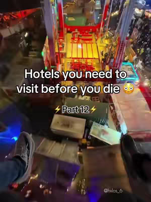 An aerial view captures an amusement park ride in operation, the camera angle giving a perspective from a seat on the ride. The ride's structure, with its red and white framework, dominates the top half of the frame, while the bottom reveals a view of the ground with assorted objects and figures. Overlaid on this scene is white text that reads "Hotels you need to visit before you die", along with the number "12" in a bolt of lightning for emphasis.