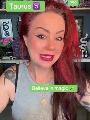 A woman with red hair is in a selfie style video. She is wearing a gray tank top and a gold necklace. Green text boxes saying "Taurus" and "Believe in magic" are overlaid on the image. The background has various items on shelves.