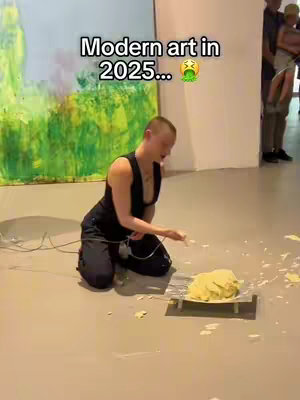 The image is a video still from an art gallery or museum. A person, likely the creator, is kneeling on the floor holding some sort of wire or cable in their right hand near a pile of yellow substance on a small wooden pallet. The background includes a large painting of green and yellow colors in a canvas frame. In the background, some people are looking at the exhibit. The text overlay reads 'Modern art in 2025...' and has an emoji.