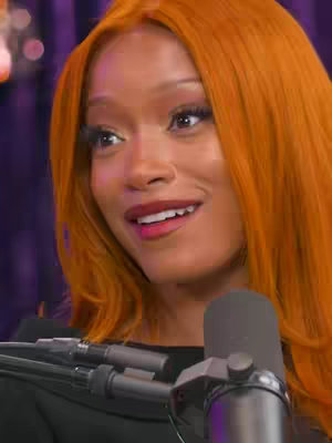 A medium close-up shot of a woman with bright orange hair. She's wearing a dark top, and a microphone partially obscures the lower part of the frame. She is looking off camera to her right smiling slightly. The background is blurred but appears to be purple with some lighting or decorative elements.