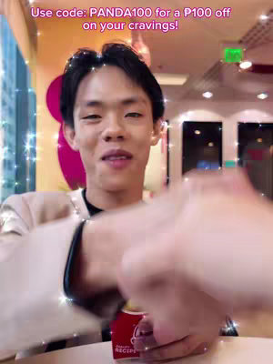 A young Asian man with black hair is smiling at the camera. He is holding a red and white product, possibly a food container, in his hands. The background is a brightly lit indoor space with some blurred elements. Text in the image promotes a discount code.