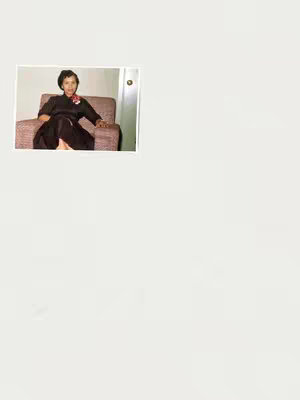 A vintage photograph framed against a white background. The photo shows a woman seated in a brown armchair positioned slightly off-center. She wears a dark, long-sleeved dress with a floral scarf at her neck. Her dark hair is neatly styled, and she is looking towards the camera. A door with a handle is partially visible in the background, and the room has cream-colored walls.