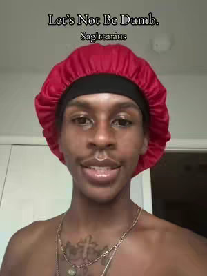 A selfie-style image featuring a person with smooth, dark skin, wearing a red satin bonnet that covers their hair. The person is looking directly at the camera with a slight smile. The person has a gold chain necklace and a tattoo on their chest. At the top of the image, a text overlay reads, 'Let's Not Be Dumb. Sagittarius.' The background appears to be a plain, light-colored wall.