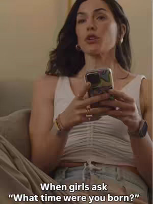 The cover image is a medium shot of a woman indoors, seated on a couch. She's holding a smartphone in front of her, seemingly looking at it. The scene appears casual, with soft lighting. The hook text 'When girls ask "What time were you born?"' is overlaid at the bottom. The layout is simple, focusing on the woman and the text, likely to grab the viewer's attention.