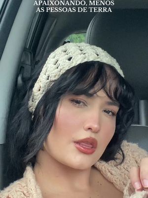 A woman in a car, looking towards the camera. She wears a cream-colored, open-weave headband and a light-colored knitted top. Her dark hair frames her face, and her mouth is slightly open. Behind her, the car interior is visible.