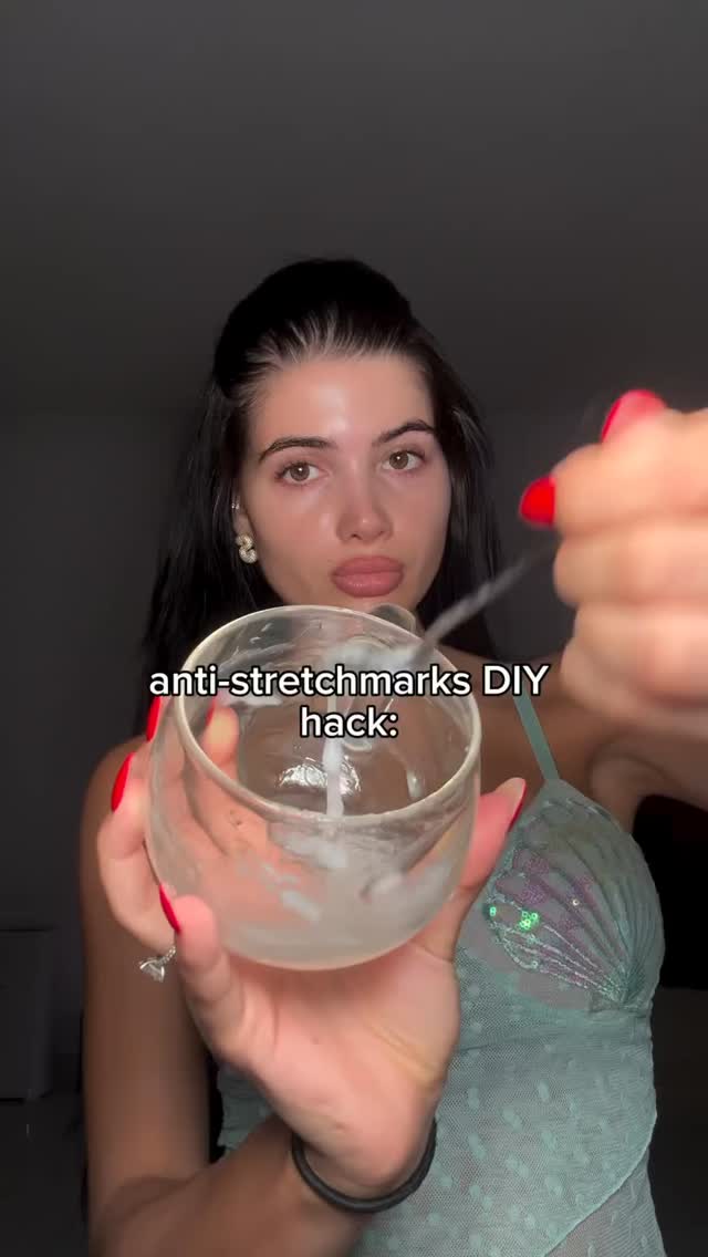 A woman with long dark hair is holding a glass with a white substance. The overlay text reads: anti-stretchmarks DIY hack. She's wearing a turquoise top, and her red-painted nails are visible as she stirs the mixture. The background is a simple, dark color.