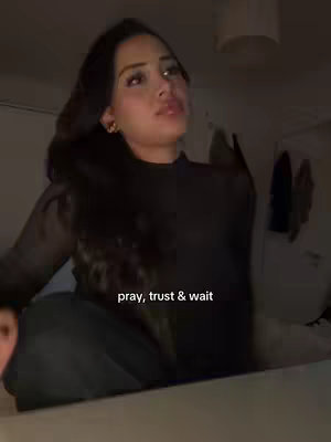 A woman is seated and looking upwards with a thoughtful expression. The image has a slightly dark, low-key lighting, with the woman's dark hair and black top contrasting against a lighter background. Text overlayed at the bottom reads "pray, trust & wait."