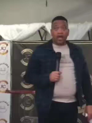A man stands facing the camera holding a microphone, with an open-mouthed surprised expression. The background has patterned fabric with banners bearing logos.