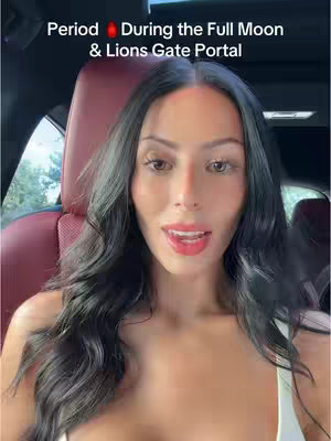 A woman with long dark hair and bright red lipstick sits in a car, facing the camera. Text overlays read, "Period During the Full Moon & Lions Gate Portal,"