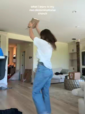 A person is posing with their arms in the air in the middle of a living room, likely capturing an outfit-of-the-day shot with a light-colored top and blue jeans. The background includes part of the interior of a house with visible furniture.