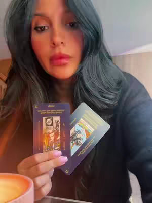 A woman with long dark hair and pink lipstick holds two tarot cards. A lit candle is in the lower left corner. The background is soft and blurred.