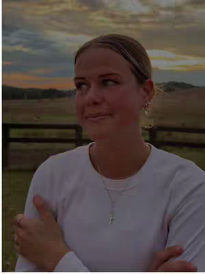 A woman gazes away from the camera with a thoughtful expression. The background shows an open grassy field under a sunset sky.