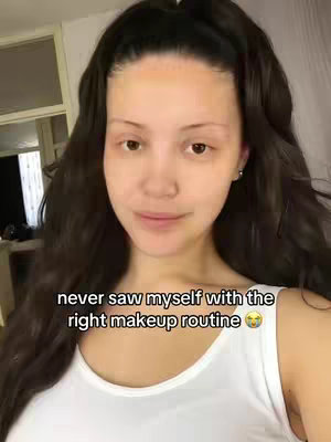 The cover image shows a woman with long wavy dark hair, gazing at the camera with a slight smile. She's wearing a white tank top. The background is softly blurred, with a glimpse of what appears to be a window or doorway. The text overlay, in a clean white font, reads, 'never saw myself with the right makeup routine' with a smiling emoji.