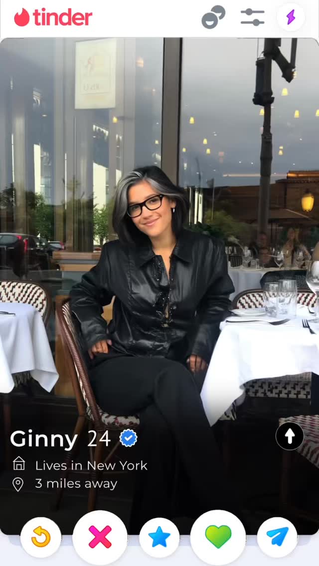 A Tinder profile screenshot with a woman seated at a restaurant table. She wears glasses and a black jacket, and the background shows a city scene with windows and tables. The interface features the Tinder logo, profile details including a name, age, location, and a set of interactive buttons.