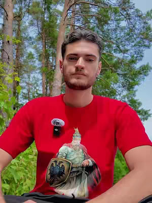 A man in a red t-shirt stands outdoors, with lush green trees in the background. He is the central figure, and the composition is focused on him. The shirt features a graphic of a figure with a shield. A microphone is attached to his shirt.