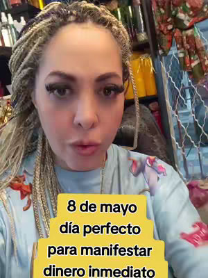 The cover image features a woman with blonde braids, looking directly at the camera with a slightly pursed lip. She's wearing a blue shirt with a design. Overlayed on her image is a multi-layered yellow text box containing the words: '8 de mayo día perfecto para manifestar dinero inmediato'. The background is blurred, showing shelves and items suggestive of a workspace.
