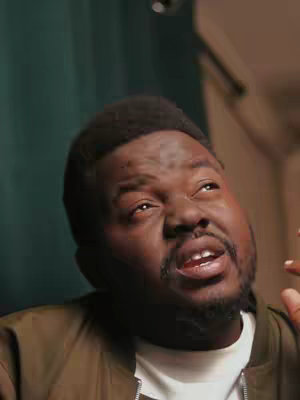 A close-up shot of a Black man looking up and to the right with a slightly perplexed expression. He is wearing a white t-shirt and a brown bomber jacket, set against a blurred background including a curtain and some neutral-toned room decor. The composition is a medium shot focusing on his face and upper body.