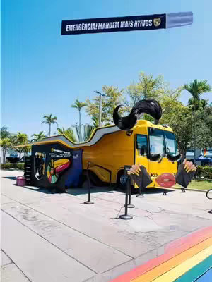 The image showcases a yellow bus with artistic modifications, set against a bright, clear sky. The bus is decorated with whimsical features, including large cartoon-like hair on top and exaggerated facial features like giant lips and eyelashes. A banner in the sky displays text in a foreign language, adding an element of international flair. A rainbow-colored path adds a vibrant touch to the foreground.
