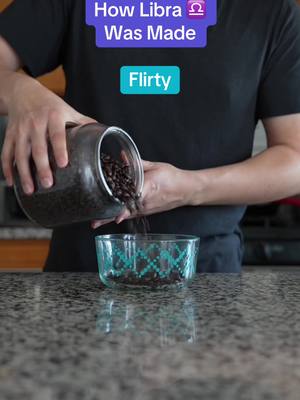 A person is pouring dark-colored beans from a glass jar into a clear glass bowl. Above the person's head, the text overlay reads 'How Libra Was Made' with the Libra zodiac symbol. Below this, centered on the image, is the text 'Flirty'. The person, dressed in a black shirt, is captured from mid-torso up, with their hands holding the jar over the bowl. The background features a kitchen with a countertop.