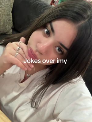 A selfie-style image featuring a young woman looking at the camera with a slightly tilted head. She is wearing a white shirt. Her left hand is near her mouth, holding up several silver rings. The background is blurred and shows a hint of a dark couch. The lighting is soft and natural. Text overlay "Jokes over imy" in white font.