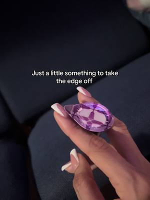 A hand with long, white-tipped nails holds a large, tear-drop shaped, purple object. It is set against a blurry dark background, possibly a piece of clothing. The object resembles a jewel.