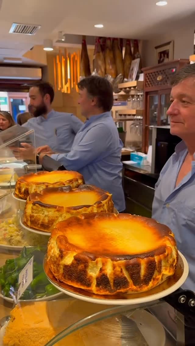 A counter displaying three Basque burnt cheesecakes with nicely browned tops, behind a glass barrier. Three men are standing behind the counter, with another customer in the background. The setting appears to be a cafe.