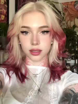 A close-up shot of a young, fair-skinned person with a pale complexion, wearing a white top. Their hair is styled with a striking color gradient, transitioning from a platinum blonde at the roots to a vibrant red towards the ends. The individual has light-colored eyes, dark eyeliner, and full, pink lips. They have a small nose ring and delicate necklaces. The background is softly focused, including foliage and possibly artwork or decor, suggesting an indoor setting. The overall composition focuses on beauty and fashion.