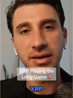 A man with short dark hair wearing white earbuds is centered in the frame, looking directly at the camera and speaking. The background appears to be a neutral, indoor setting. Overlaid on the image is a rectangular text box containing the phrase "XRP Playing the Long Game", with a target emoji next to it, and the text "XRP" in a blue box at the bottom.